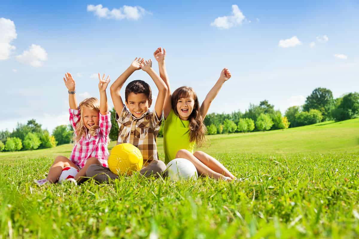 Portrait of three happy kids, boy and girls sitting in the grass in park with lifted hands and holding sport balls