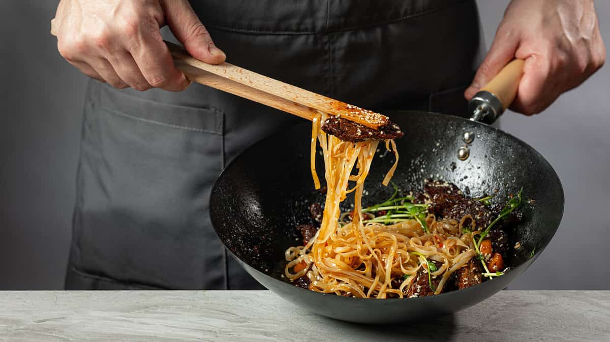 Asian fast food rice clear noodles cooking with fried meat and spicy vegetables by cook chef hands in wok pan. Close up food recipe concept.