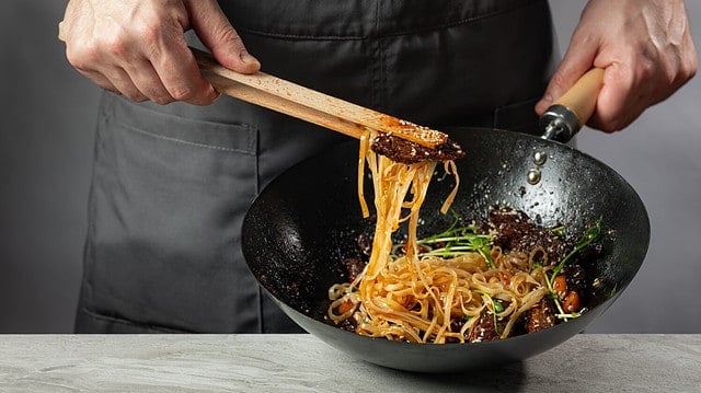 Asian fast food rice clear noodles cooking with fried meat and spicy vegetables by cook chef hands in wok pan. Close up food recipe concept.