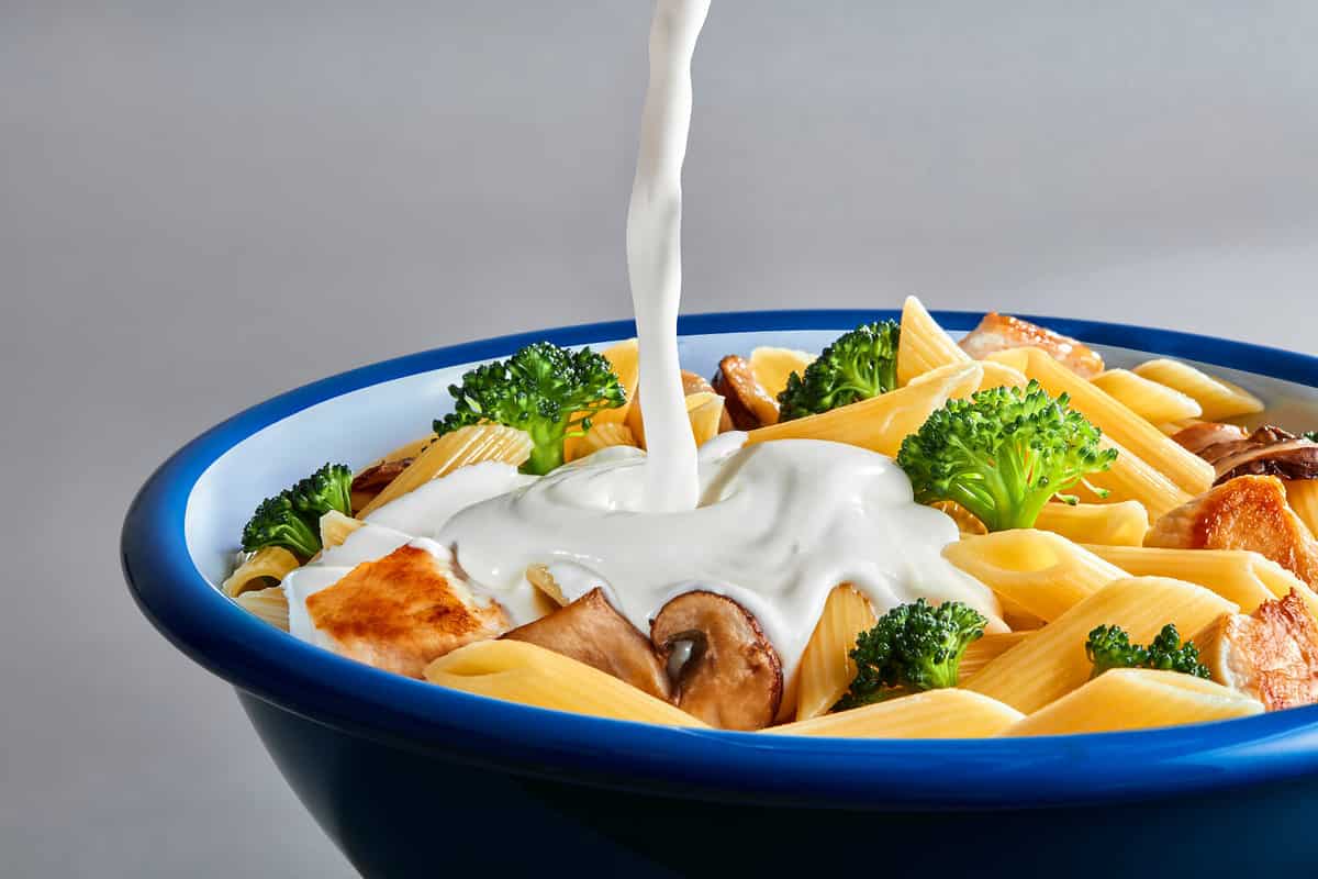 Pouring white cream sauce over plate of pasta.