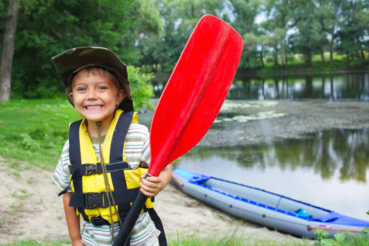 Portrait of happy young boy holding paddle near a kayak on the river, enjoying a lovely summer day