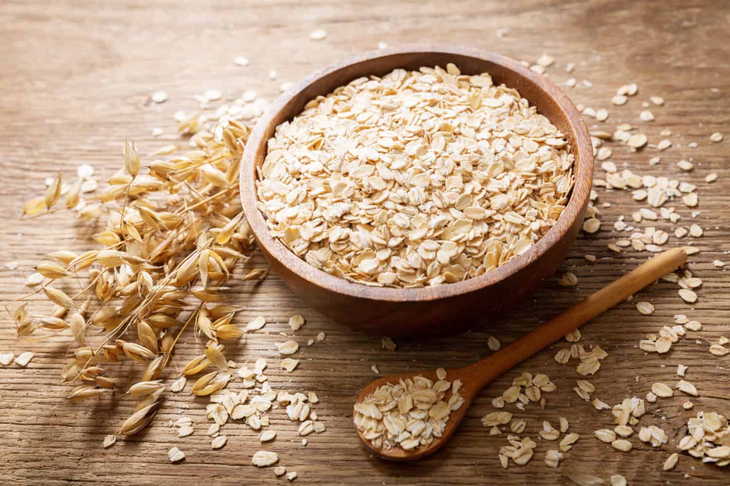 A bowl of oats with ears of oats, and flakes of oatmeal scattered on a wooden table.