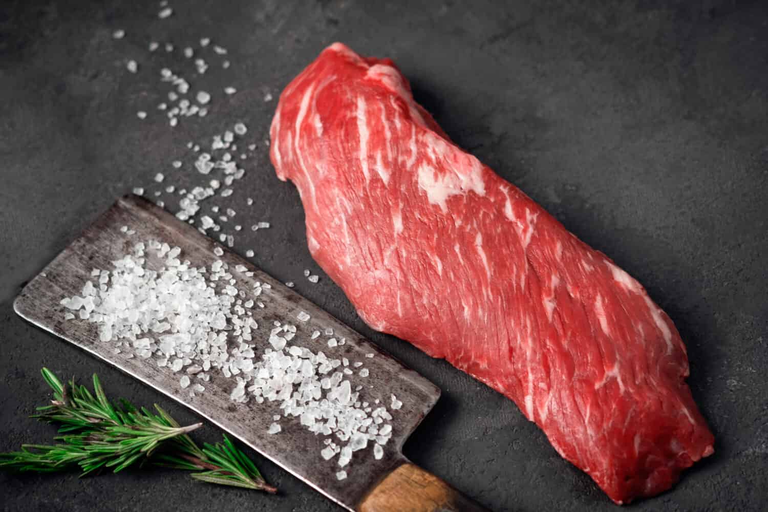 Raw tenderloin of beef or skirt steak with a hatchet for meat, salt and rosemary on a dark stone background, close up