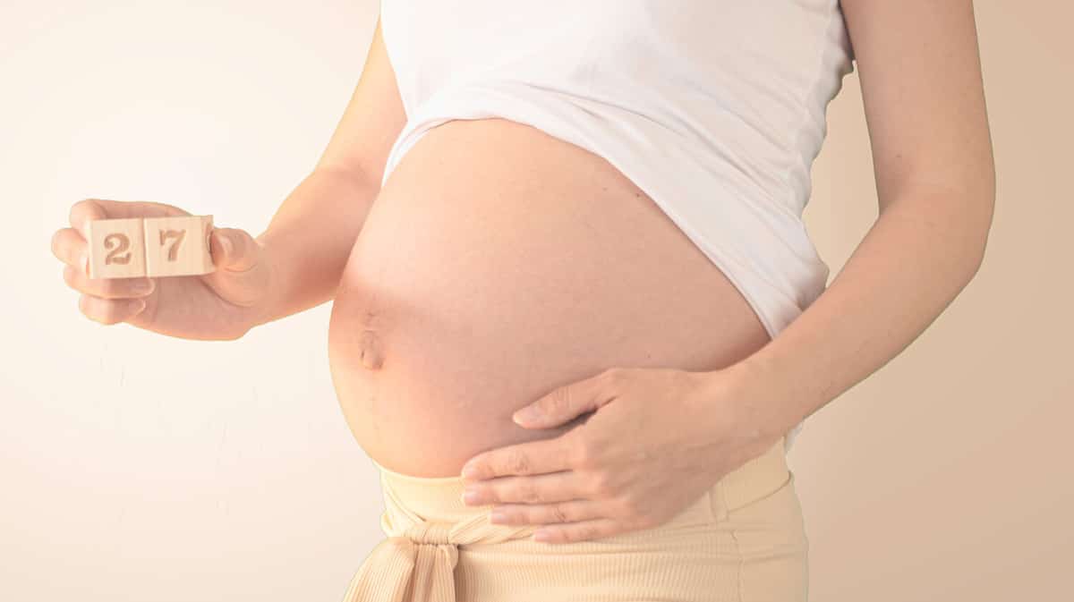 Pregnant young woman with pregnancy week number next to her belly. Photos of belly growth at 27 weeks pregnancy. Healthy pregnancy diet and fetal development.