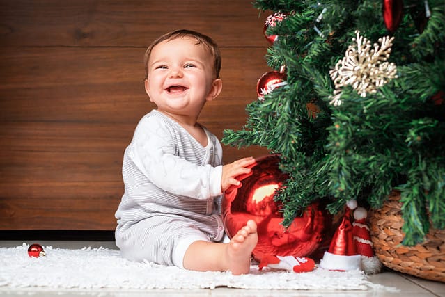 cute child with christmas tree. happy baby sitting near a fir tree and holding a christmas ornament and smiling