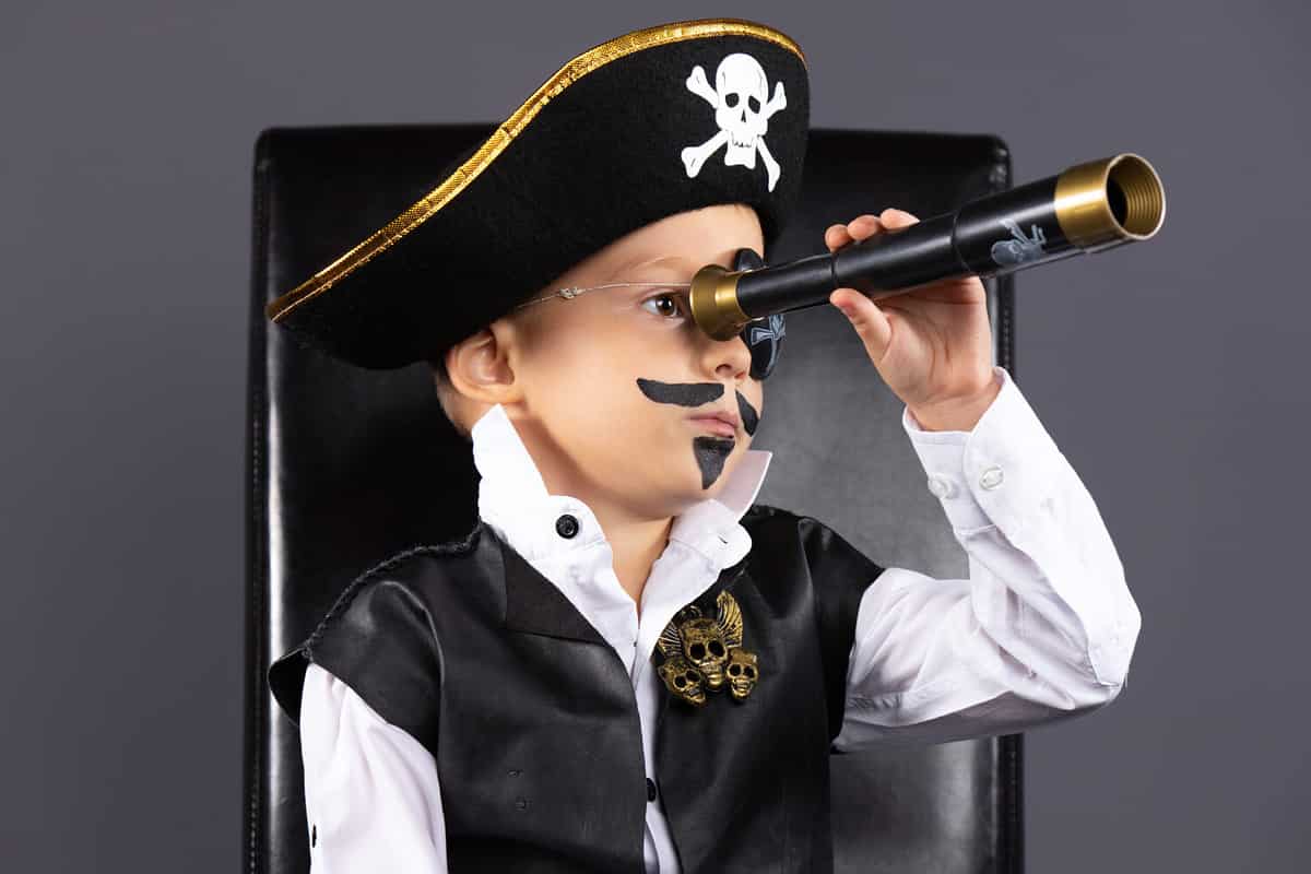 Awesome caucasian kindergarden boy with face painted sitting on the black leather chair. He is disguised as a pirate scanning the horizon through . Halloween concept isolated on white background