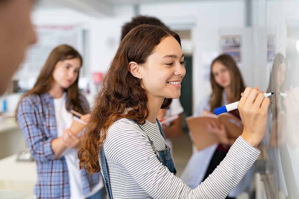 Happy college student writing equation on white board in class. Satisfied young girl solving math problem on whiteboard with classmates in background watching her. Proud high school student writing.