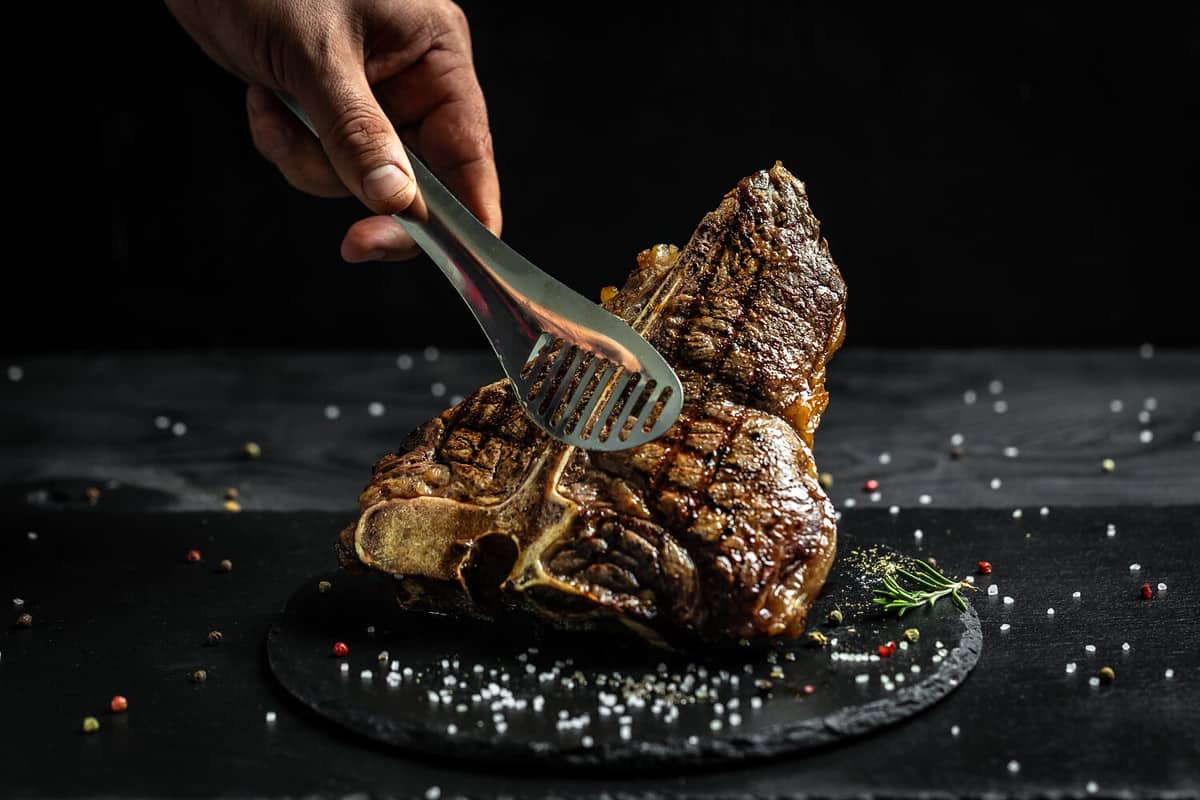 chef hand holding steak meat tongs. large piece of fresh beef meat prepared on a grill. Medium rare Grilled T-Bone Steak, Barbecue aged wagyu porterhouse.