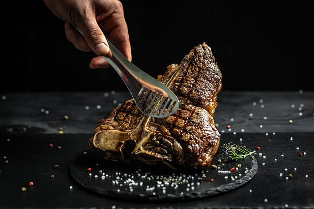chef hand holding steak meat tongs. large piece of fresh beef meat prepared on a grill. Medium rare Grilled T-Bone Steak, Barbecue aged wagyu porterhouse.