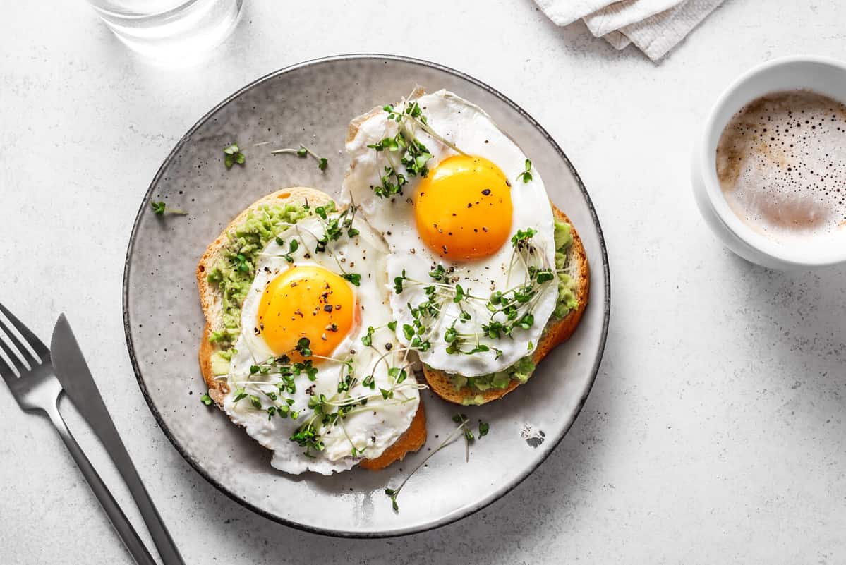 Avocado Egg Sandwiches and coffee for healthy breakfast. Whole grain toasts with mashed avocado, fried eggs and organic microgreens on white table.