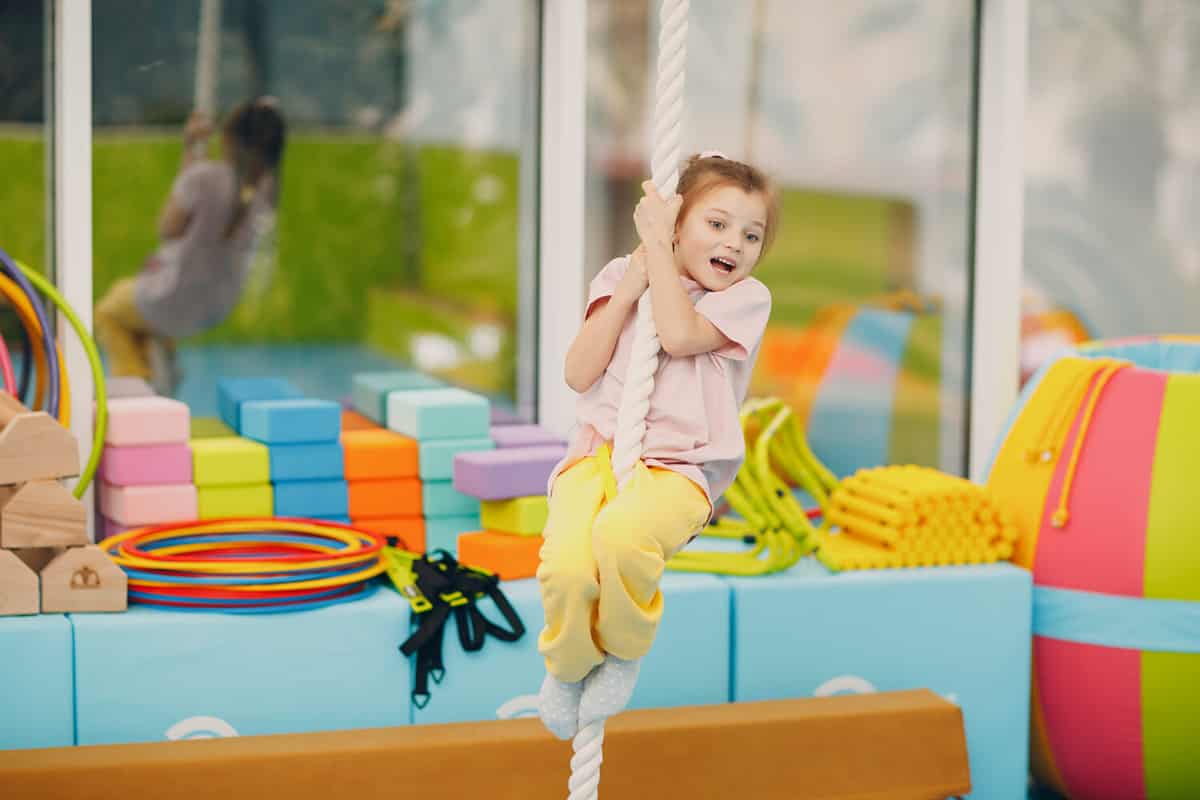 Kid girl doing exercises climbing tightrope in gym at kindergarten or elementary school. Children sport and fitness concept.