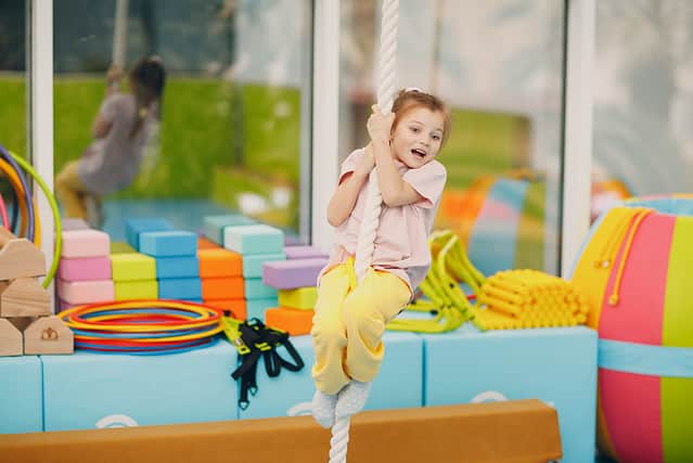 Kid girl doing exercises climbing tightrope in gym at kindergarten or elementary school. Children sport and fitness concept.