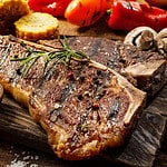 Close up of a succulent tender grilled porterhouse steak seasoned with pepper and rosemary on a wooden board with fresh halved tomatoes, mushrooms, corncobs and bell peppers