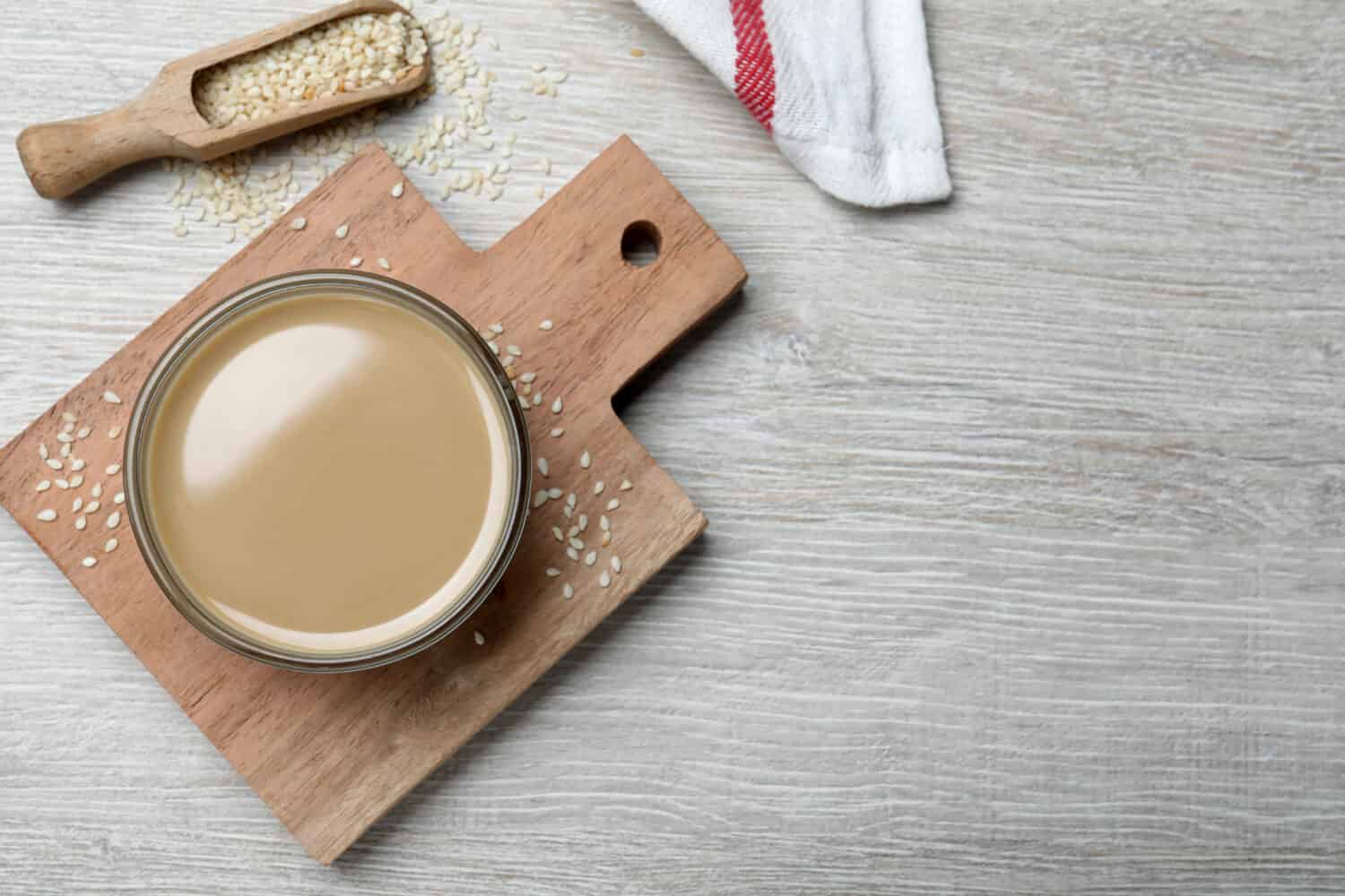 Tasty sesame paste, seeds and scoop on white wooden table, flat lay. Space for text