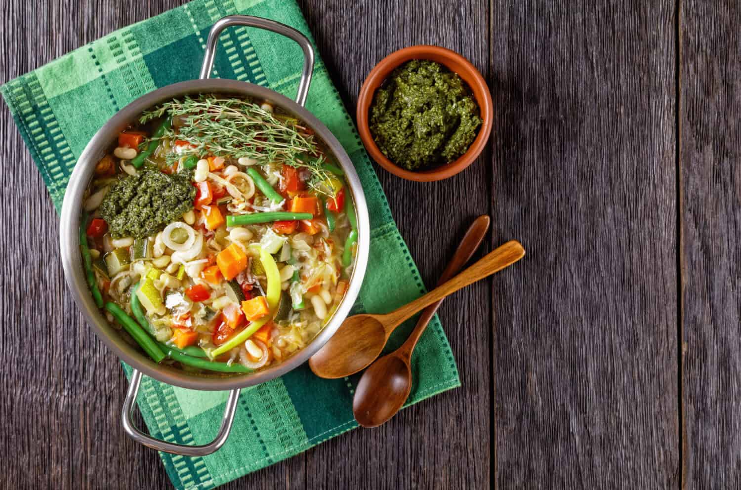 Soupe au Pistou, french vegetable bean soup with basil sauce pesto and herbs in a metal pot on a dark rustic wooden table with wooden spoon, flat lay, free space
