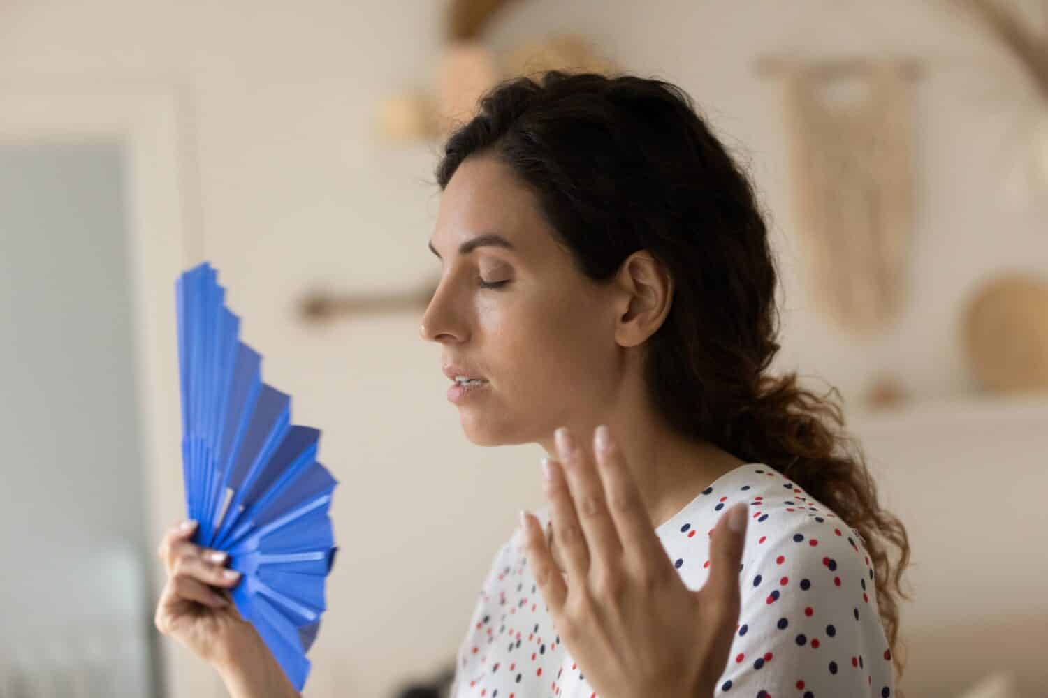 Exhausted overheated sweaty woman suffering from heat at home without conditioner, trying to cool too hot air with handheld fan in summer day, feeling unwell du to weather, humidity, air temperature