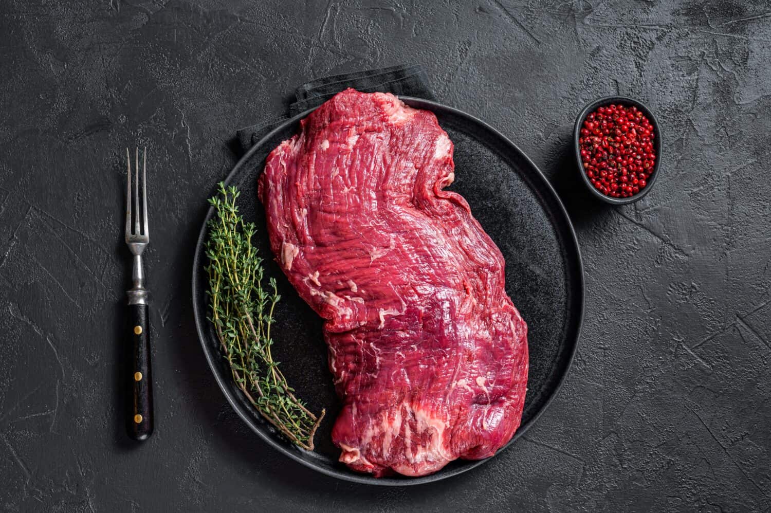 Flank or flap raw beef steak on plate with thyme. Black background. Top view.
