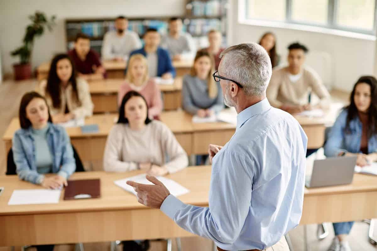 Back view of mature professor giving lecture to large group of college students in the classroom.