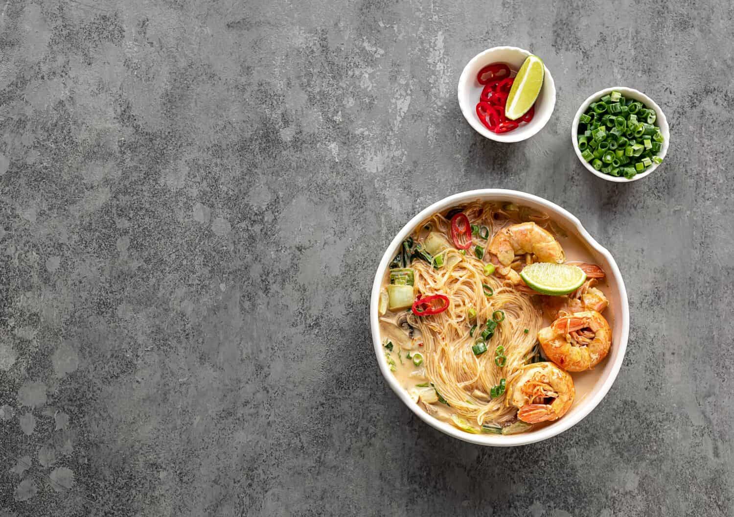 Bowl of curry laksa, a spicy glass noodle dish popular in Southeast Asia with prawns, bok choy, lime, ginger, and chili. Most variations of laksa are prepared with a rich and spicy coconut soup.