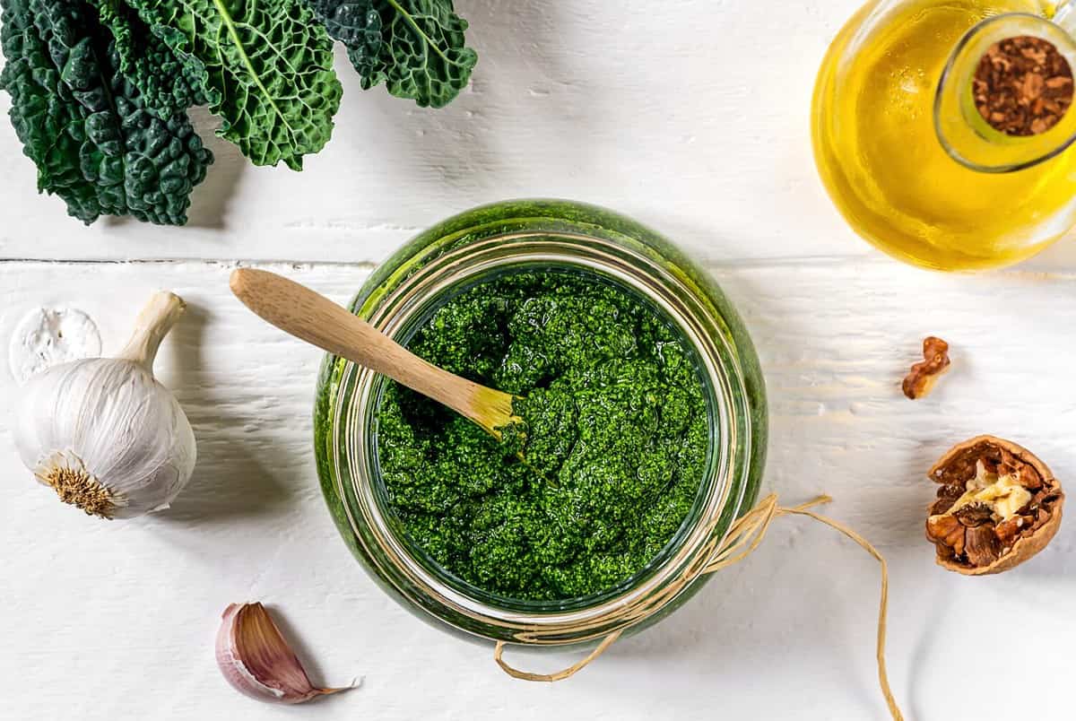 Homemade kale pesto sauce served in a glass bowl over white wooden surface with basic ingredients - kale leaves, walnuts, parmesan, garlic and olive oil