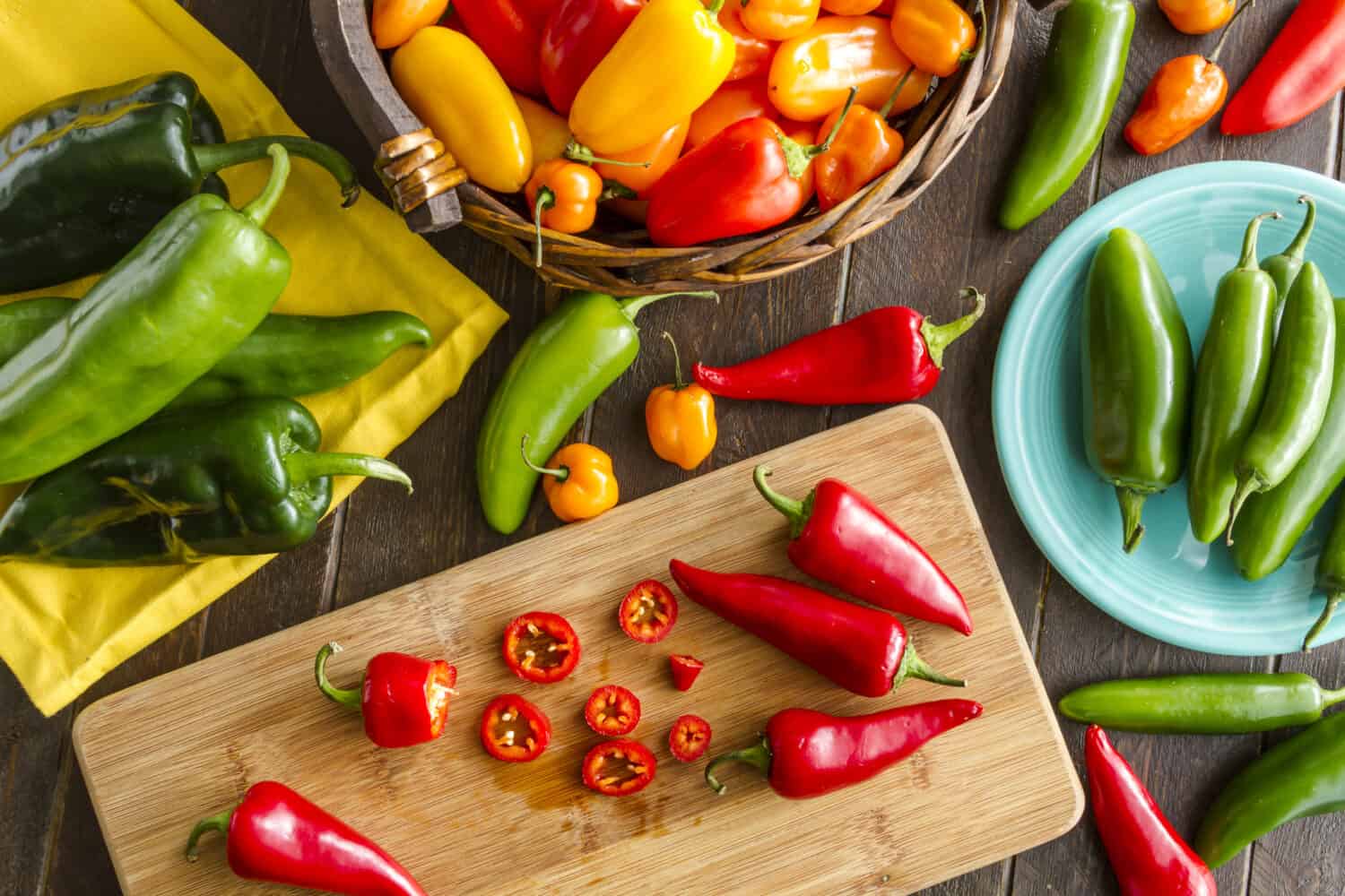 Assorted colorful varieties of hot and sweet peppers sitting on table with cutting board, yellowe napkin and blue plate