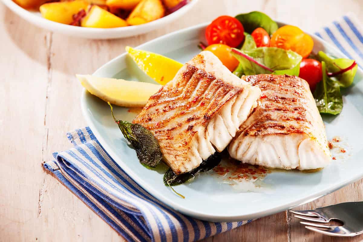 To portions of fresh grilled pollock or coalfish served with colorful salad and slices of lemon, close up high angle view