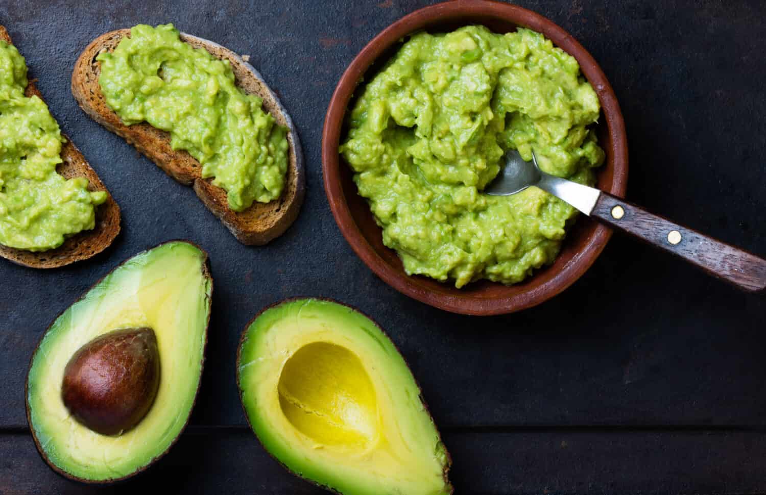 Traditional latinamerican mexican sauce guacamole in clay bowl, cut half avocado and avocado sandwiches on dark background. Top view