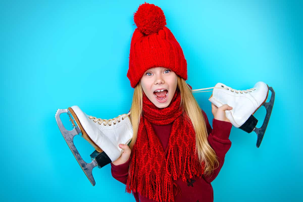 Cheerful little girl in warm sweater and hat holding figure skates. Blue background.