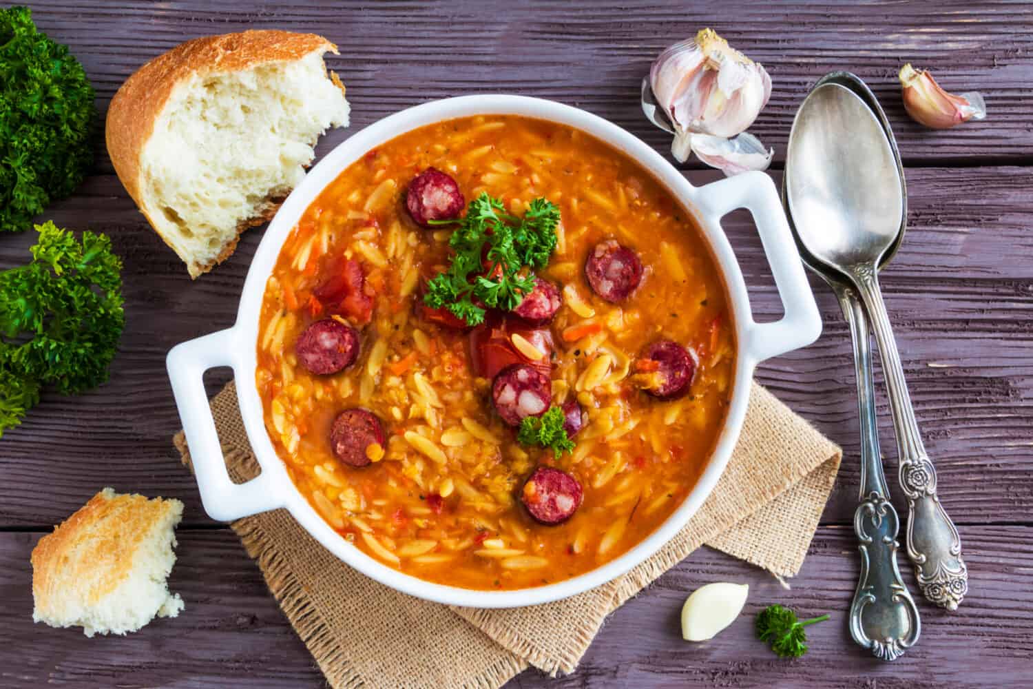 Tomato soup with orzo and smoked sausages in white casserole on wooden rustic table. Fresh bread and parsley, vintage spoon, top view.