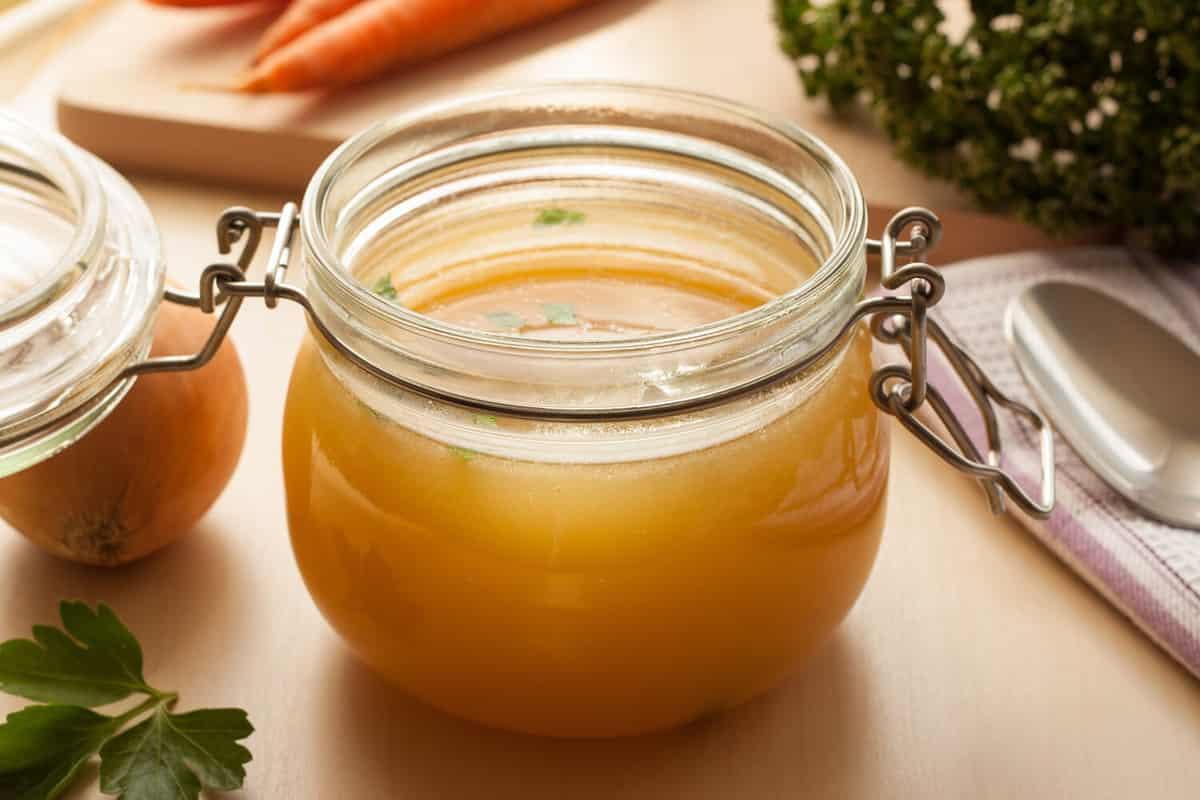 Bone broth made from chicken in a glass jar, with carrots, onions, and parsley in the background