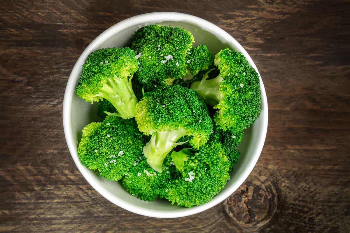 A bowl of cooked green broccoli with sea salt, shot from above on a dark wooden rustic texture with a place for text