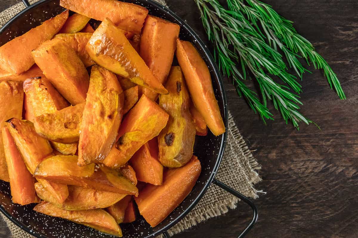 An overhead photo of roasted sweet potatoes in a pan, shot from above on dark rustic wooden textures with rosemary branches, with a place for text