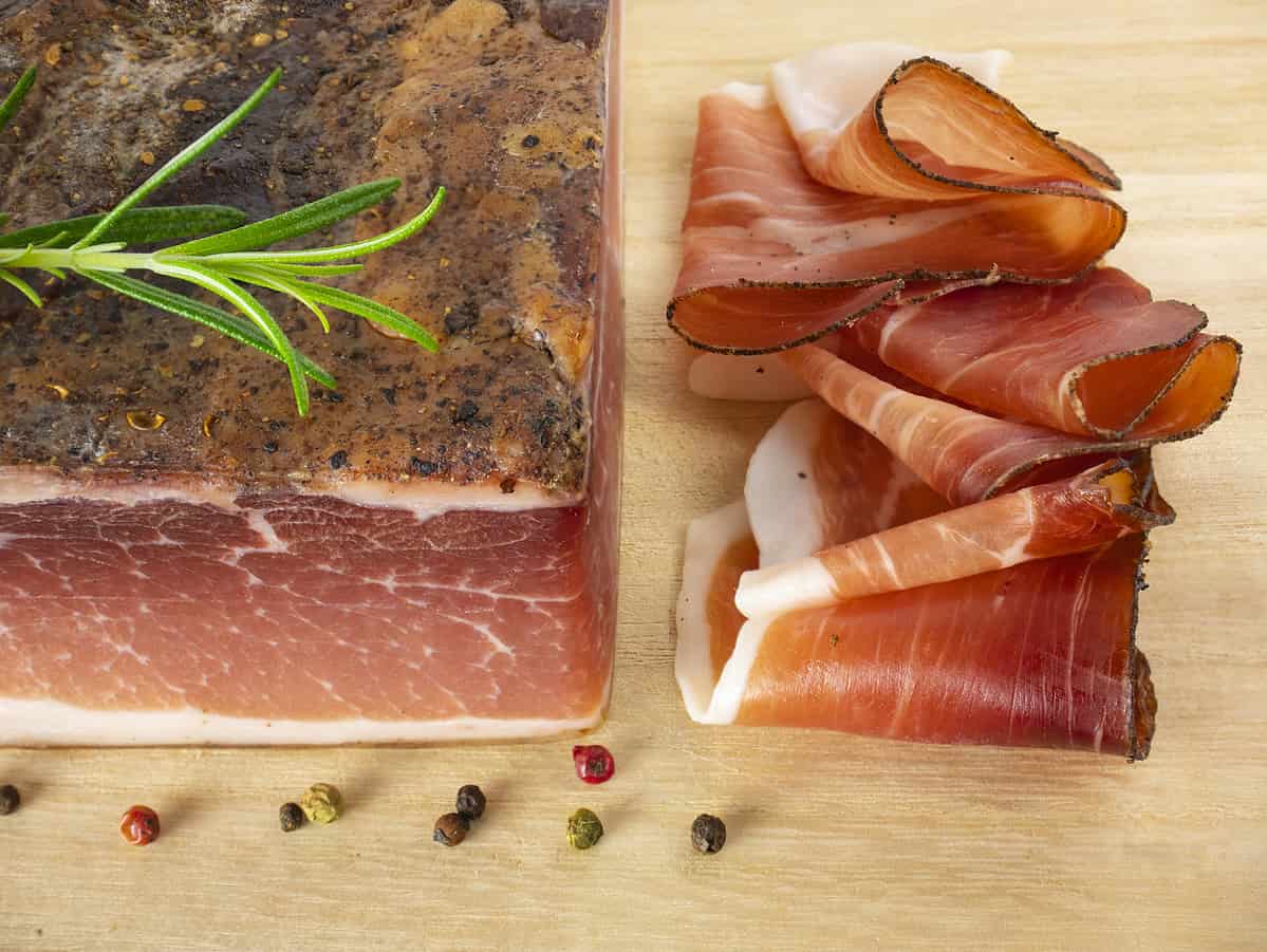 Speck with slices on the cutting board. Typical South Tyrolean smoked bacon. Sliced raw ham. Dry cured meat. Traditional cold cuts, Italian speck with rosemary and pepper. Still life food.