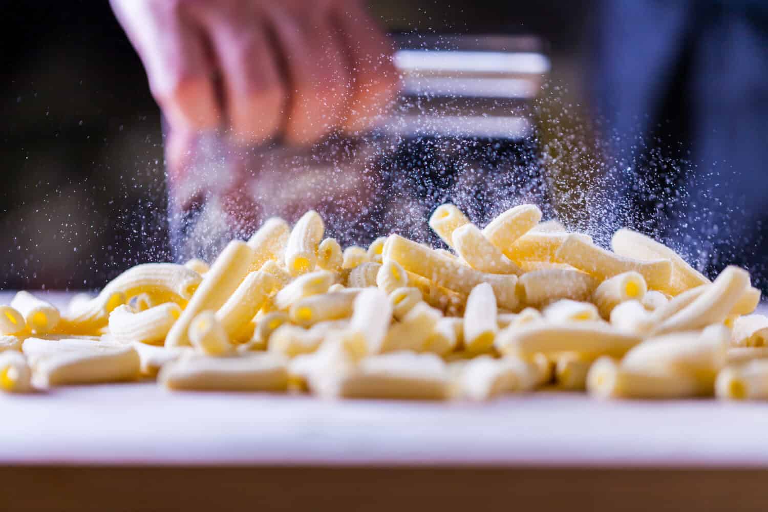 Flour falling over pasta. Falling penne pasta. Rigatoni pasta with flour powder particle falling. Rigatoni Penne Lisce Tortiglioni Doppia Rigatura. Home made rigatoni pasta on the table with flour.