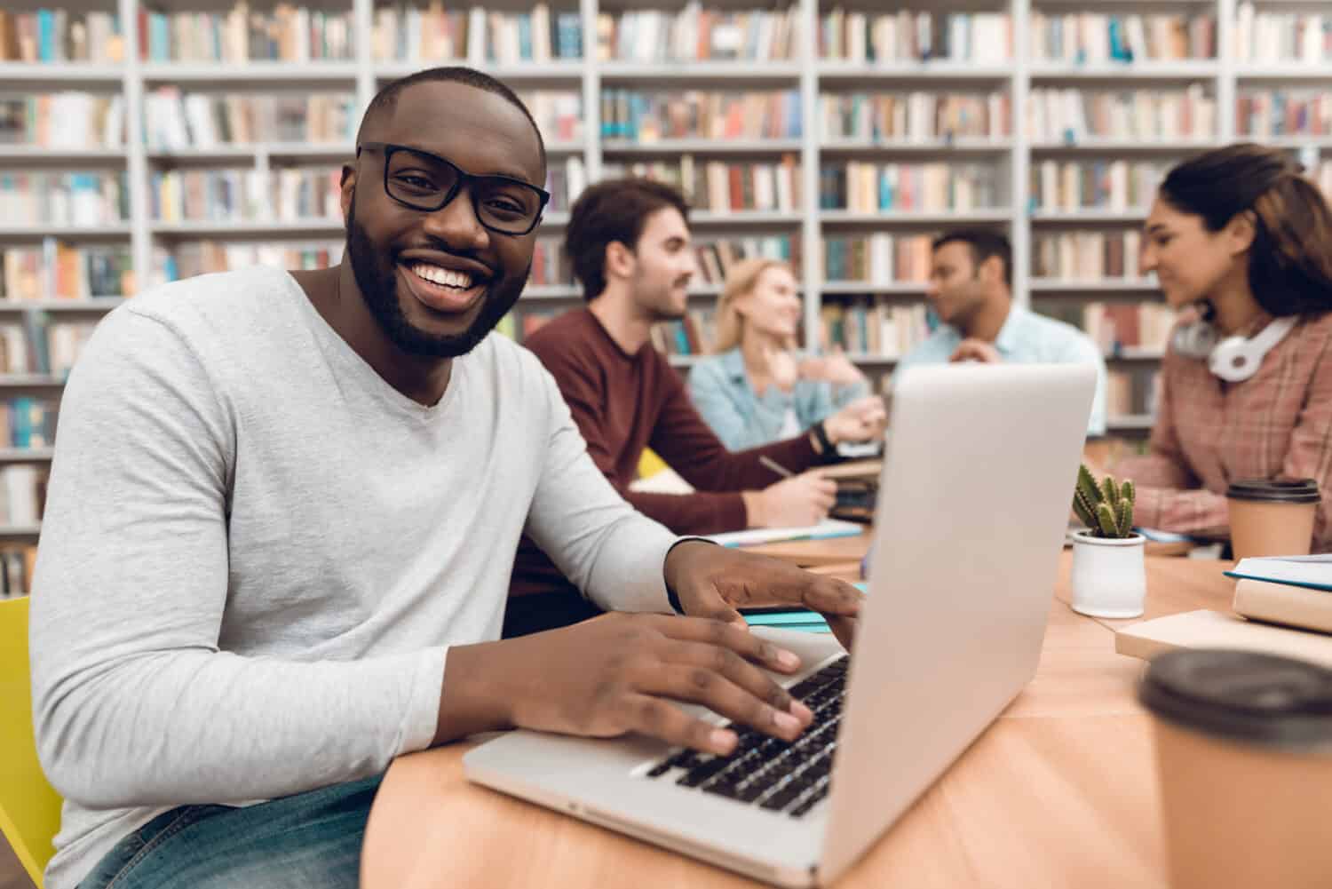 Group of ethnic multicultural students sitting at table in library. Black guy on laptop.