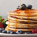 Close-up delicious pancakes, with fresh blueberries, strawberries and maple syrup on a light background. With copy space. Sweet maple syrup flows from a stack of pancake