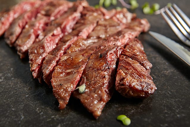 Thick Slices of Hot Grilled Whole Machete Steak or Skirt Steak on Black Stone Background. Fresh Juicy Medium Rare Beef Grillsteak. Barbecue Meat Top View Close Up