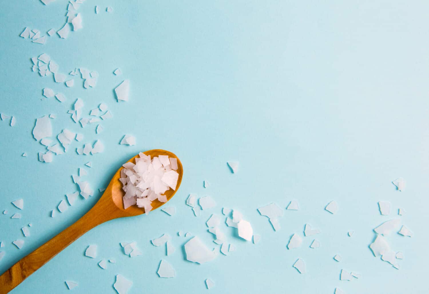 Magnesium Chloride Flakes scattered around brown wooden spoon on blue background. For making foot bath, taking a magnesium-rich bath allows full body exposure to a concentrated solution of magnesium.