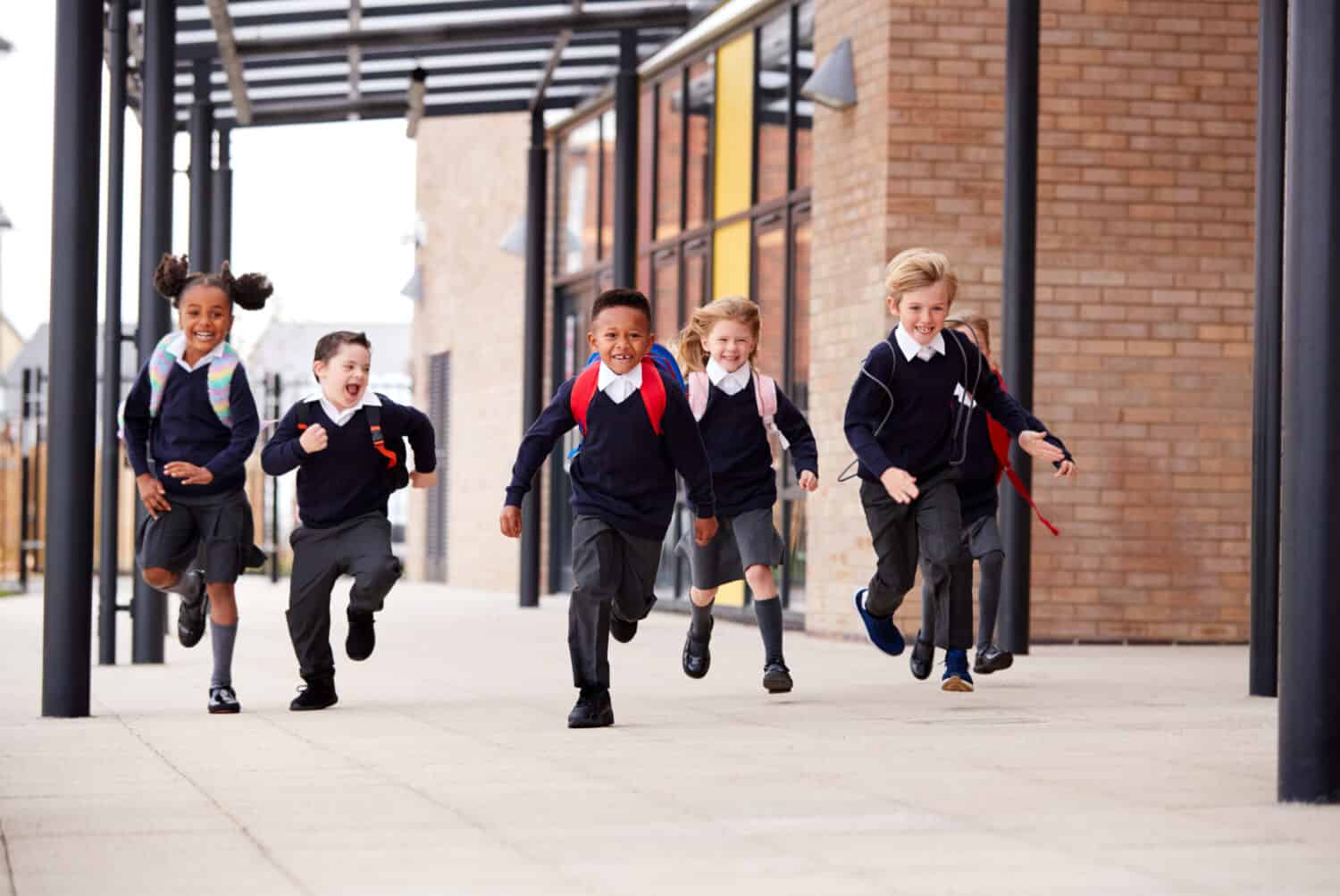 Primary school kids, wearing school uniforms and backpacks, running on a walkway outside their school building, front view