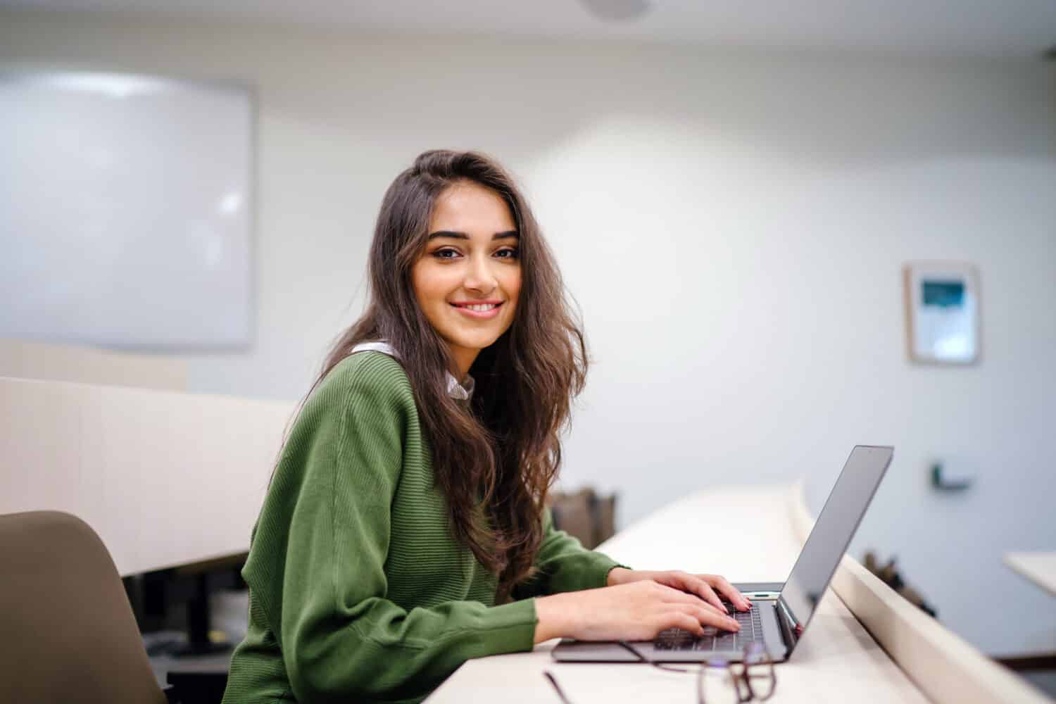 A university student looking at the camera smiling while working on her laptop.