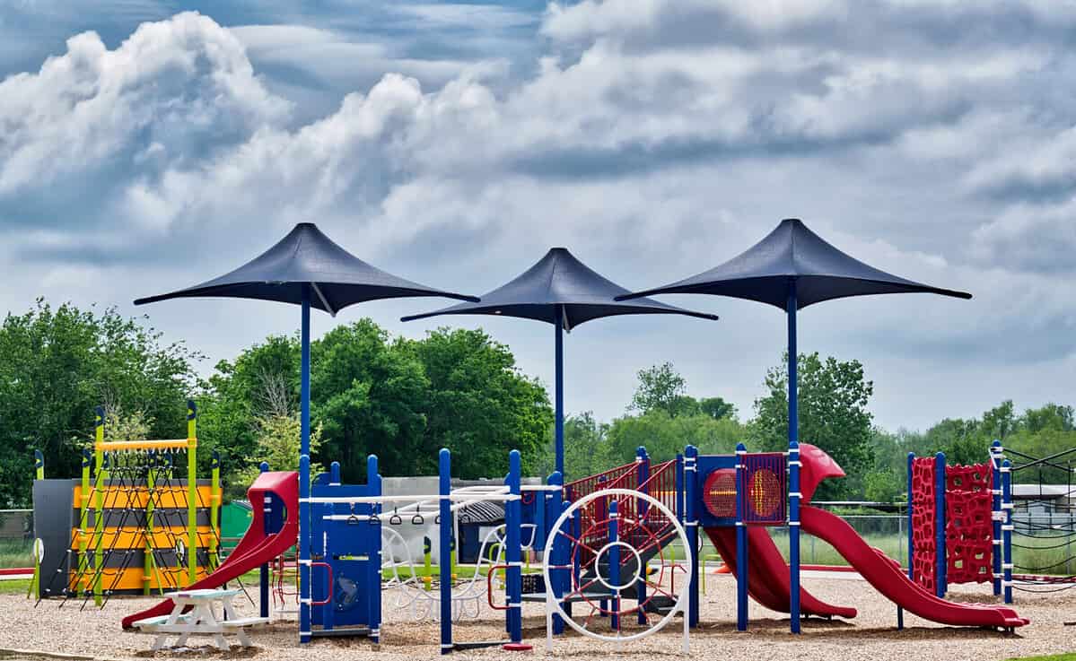 A public school playground sits deserted during the coronavirus quarantine period in Houston, TX. Three shade umbrellas stand out from the colorful equipment against a dramatic cloudy sky.