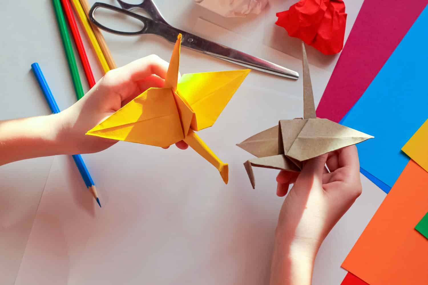 Children’s hands doing origami crane from yellow paper on white background with various school supplies. Step-by-step tutorial of origami. Step 18. Concept of children's creativity, back to school.