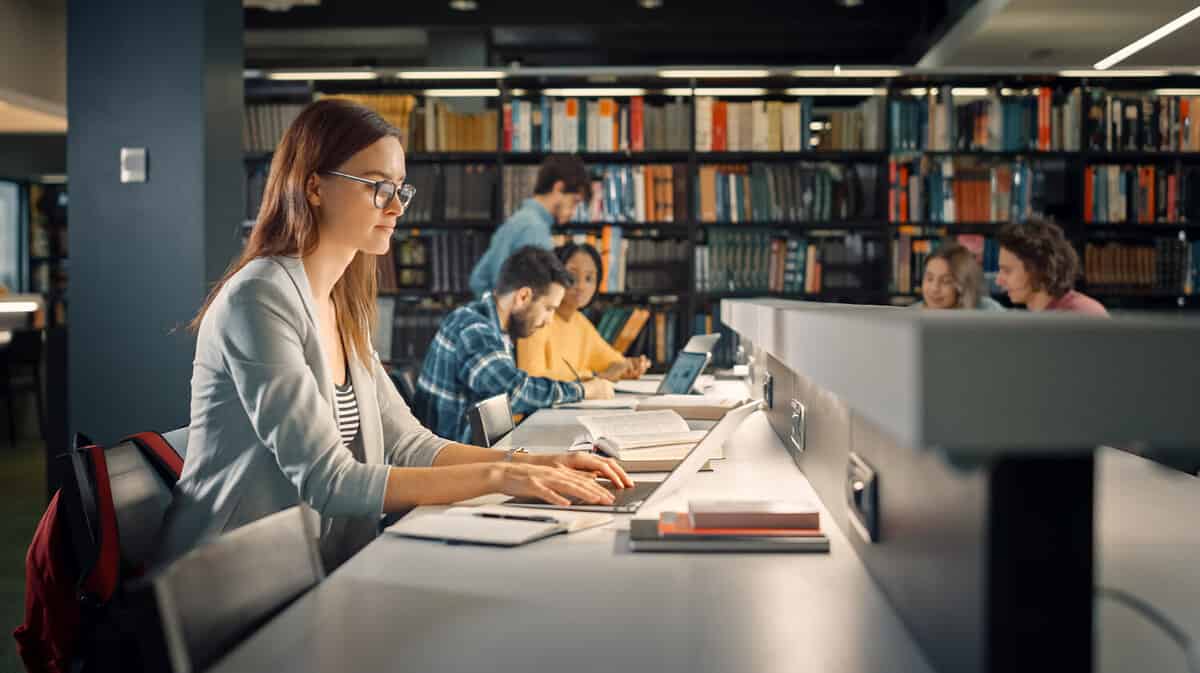 University Library: Talented Caucasian Girl Sitting at the Desk, Uses Laptop, Writes Notes for the Paper, Essay, Study for Class Assignment. Diverse Group of Students Learning, Studying for Exams.