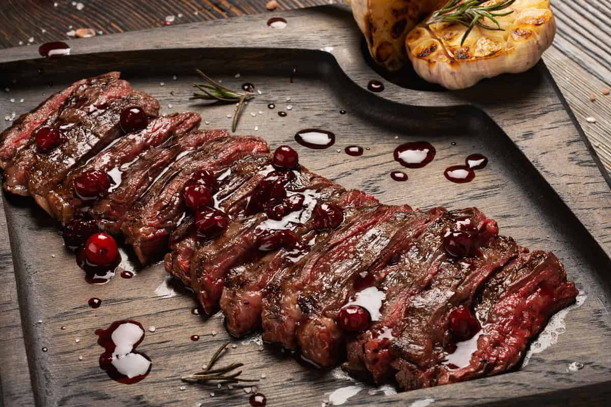 Prime Black Angus Skirt steak with cranberry sauce and grilled garlic on wooden board. Medium Rare degree of steak doneness.