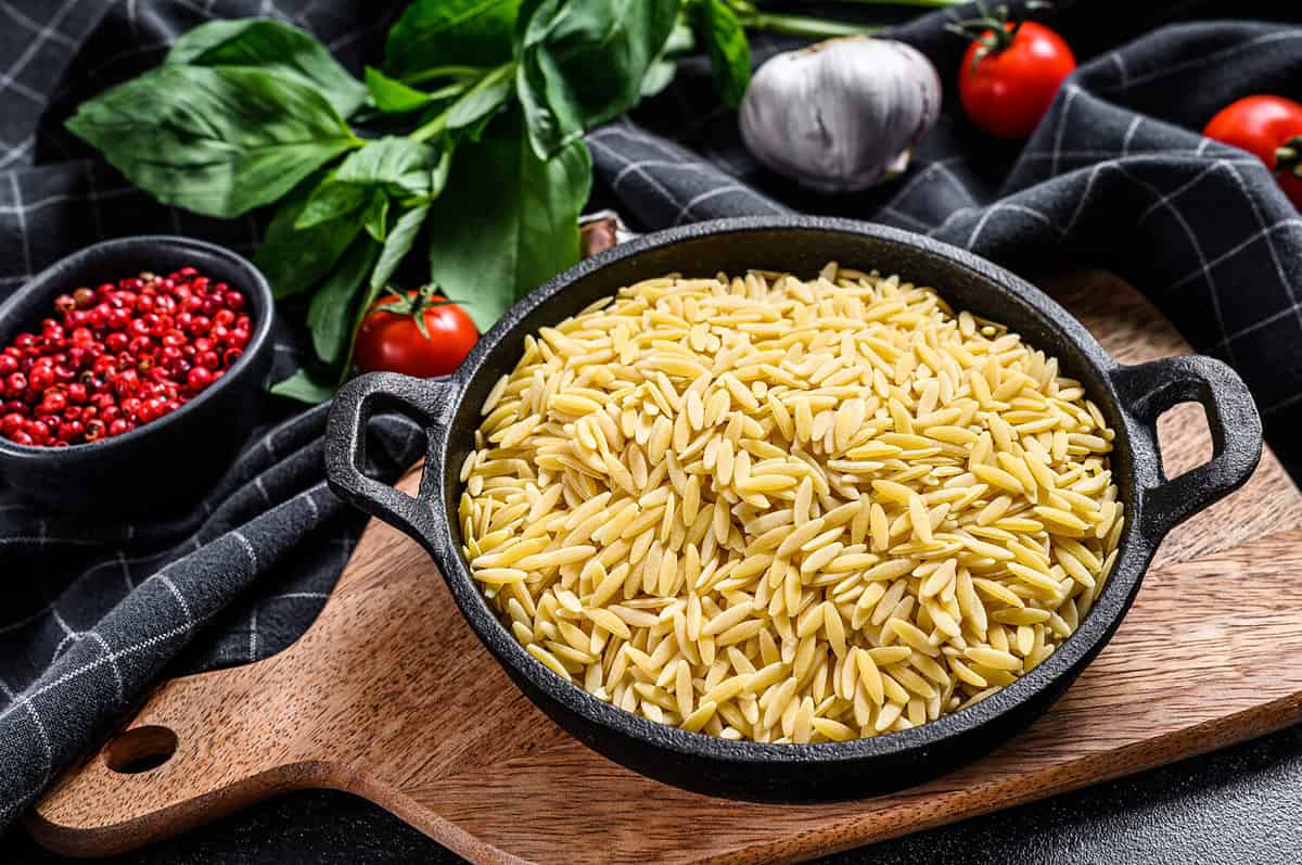 A bowl of uncooked Orzo pasta with Basil leaves, tomatoes and garlic. Black background. Top view.