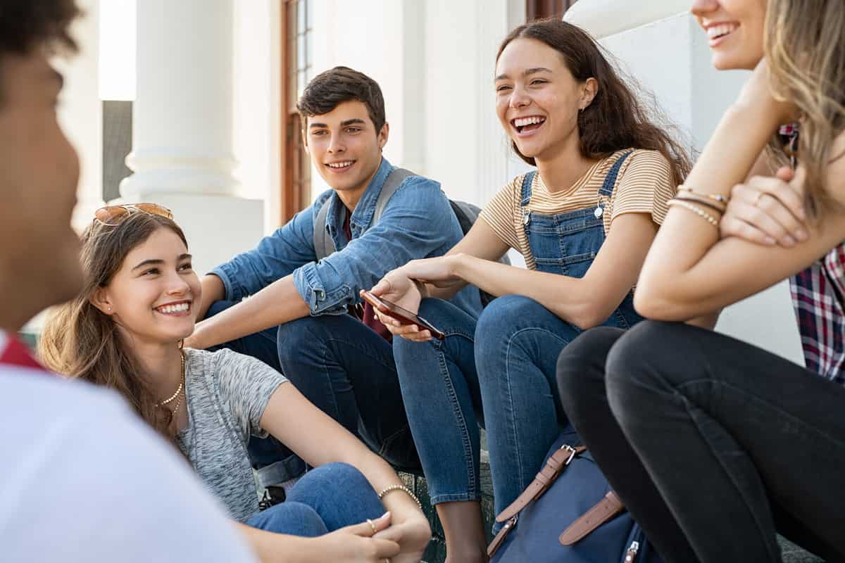 Group of happy young friends sitting in college campus and talking. Cheerful group of smiling girls and guys feeling relaxed after university exam. Excited millenials laughing and having fun outdoor.