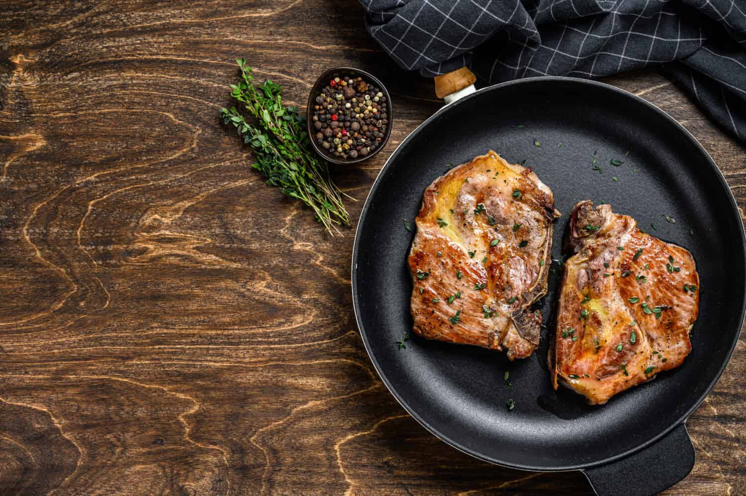 Roasted pork steaks in frying pan from neck meat. Dark wooden background. Top view. Copy space.