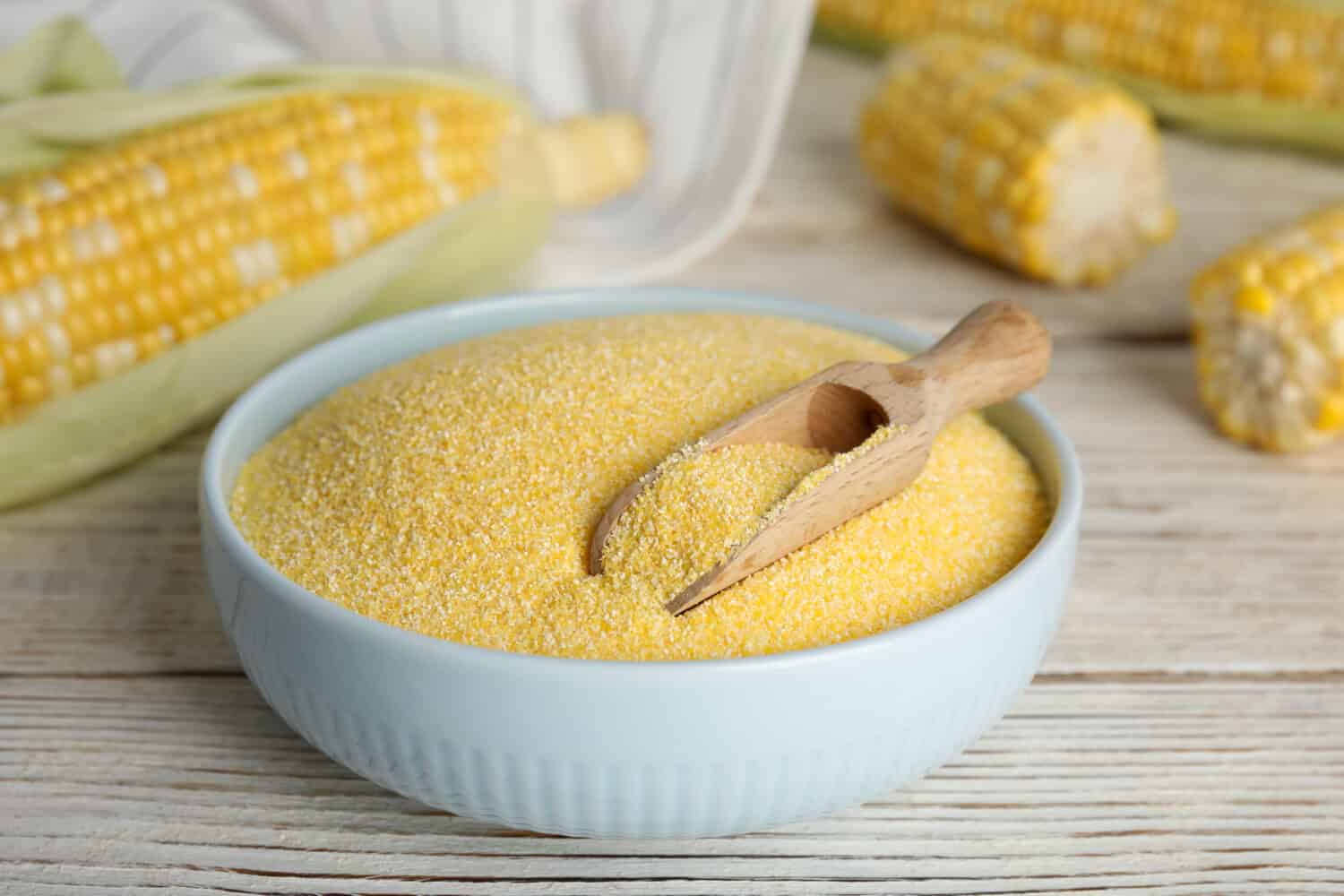 Cornmeal in bowl and fresh cobs on white wooden table