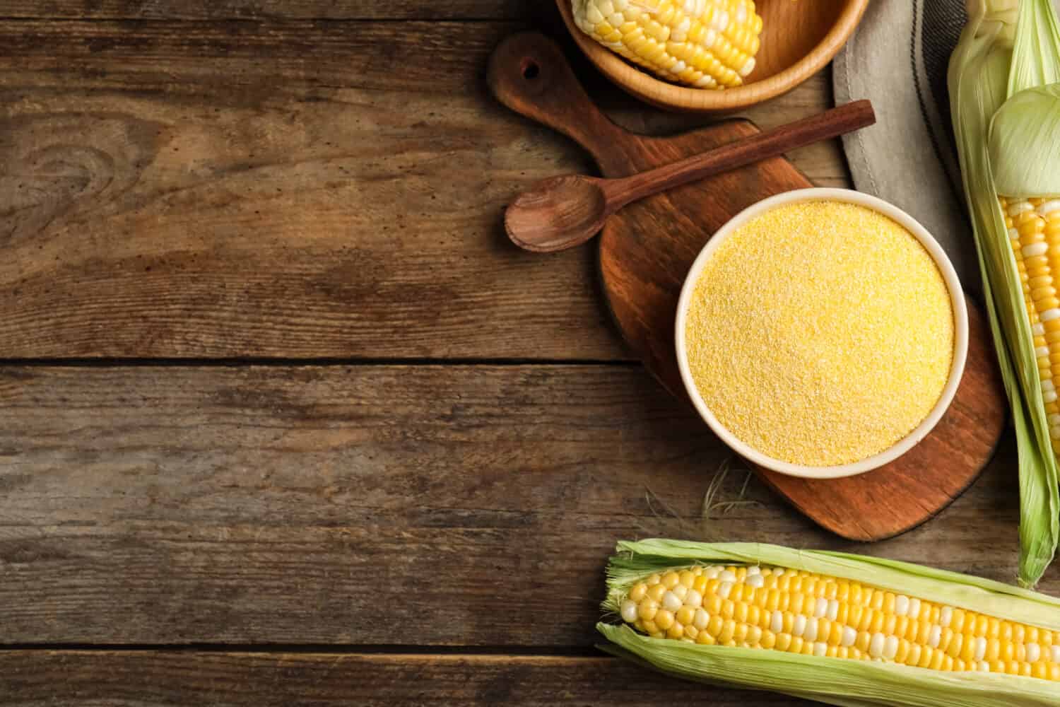 Cornmeal in bowl and fresh cobs on wooden table, flat lay. Space for text