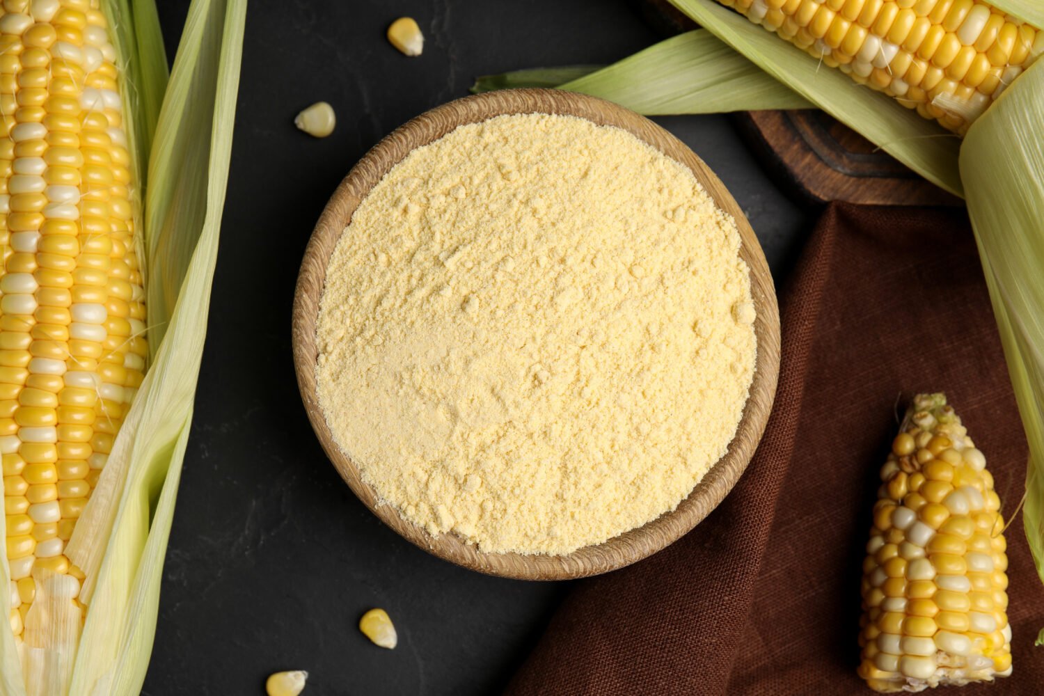 Corn flour in bowl and fresh cobs on black table, flat lay